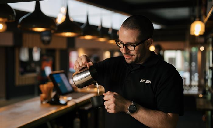 A man stands behind a bar as he pours milk out of a jug into a coffee cup. He wears a watch, glasses and a dark t-shirt with a logo that reads 'MWLDAN